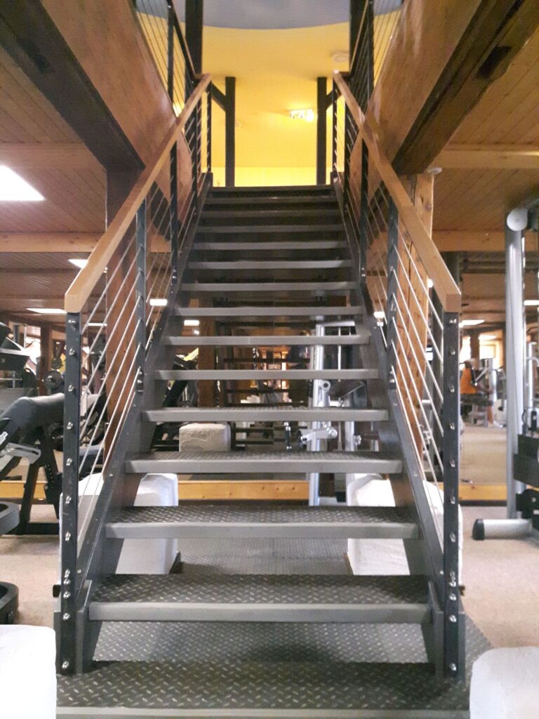 Gerade Stahltreppe mit Riffelblechstufen und Holzhandlauf in einem Fitnessstudio